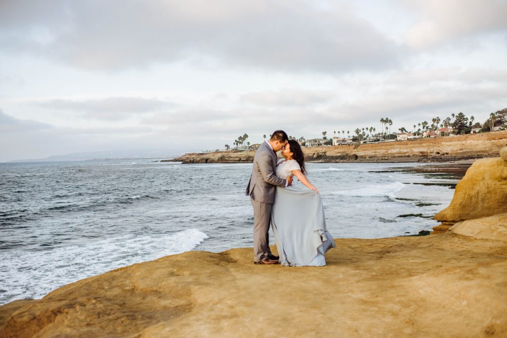 Groom holding his bride as her beaitufl light blue skirt blows in the wind at Sunset Cliffs in San Diego, Ca